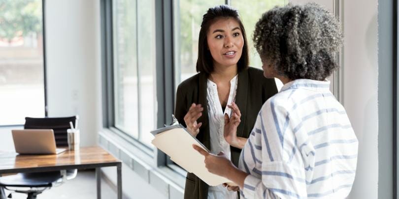 Two women in a modern office environment, one with Asian features giving feedback to an African-American colleague holding a clipboard. They are discussing strategies to help staff grow, emphasizing a collaborative atmosphere. Desk in the background with a laptop and the Xseed Lead logo subtly integrated.