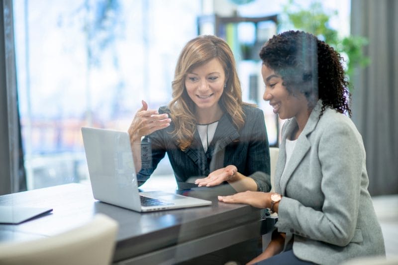 Two professional women engaging in a discussion over a laptop in a bright office setting.