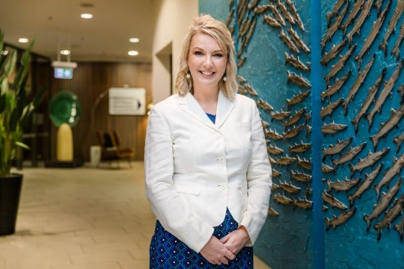 A professional woman in a white blazer and patterned skirt stands smiling in a modern office lobby with a decorative blue wall sculpture behind her.