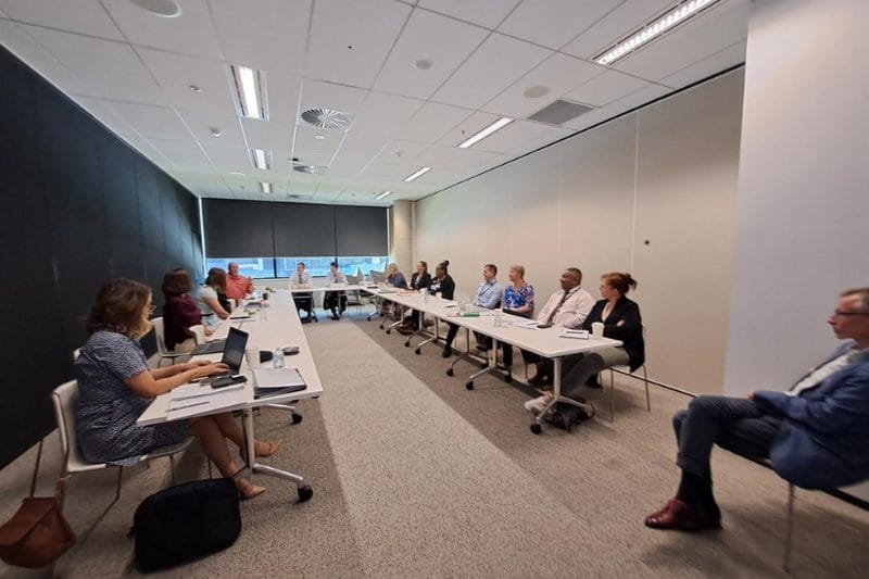 Modern training session at Xseed Lead’s Training and Capacity Development program, showing participants of various ethnicities seated along a long table in a sleek, well-lit conference room. They are actively engaged, some looking towards the speaker, others at their laptops, emphasizing a collaborative and inclusive learning environment.
