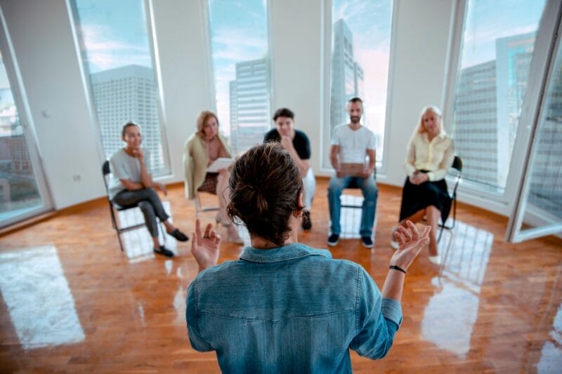 A diverse group of professionals participating in a workshop titled "How to Tell Someone They Were Not Successful in a Promotion Round" led by a facilitator in a bright office with large windows overlooking a city skyline.