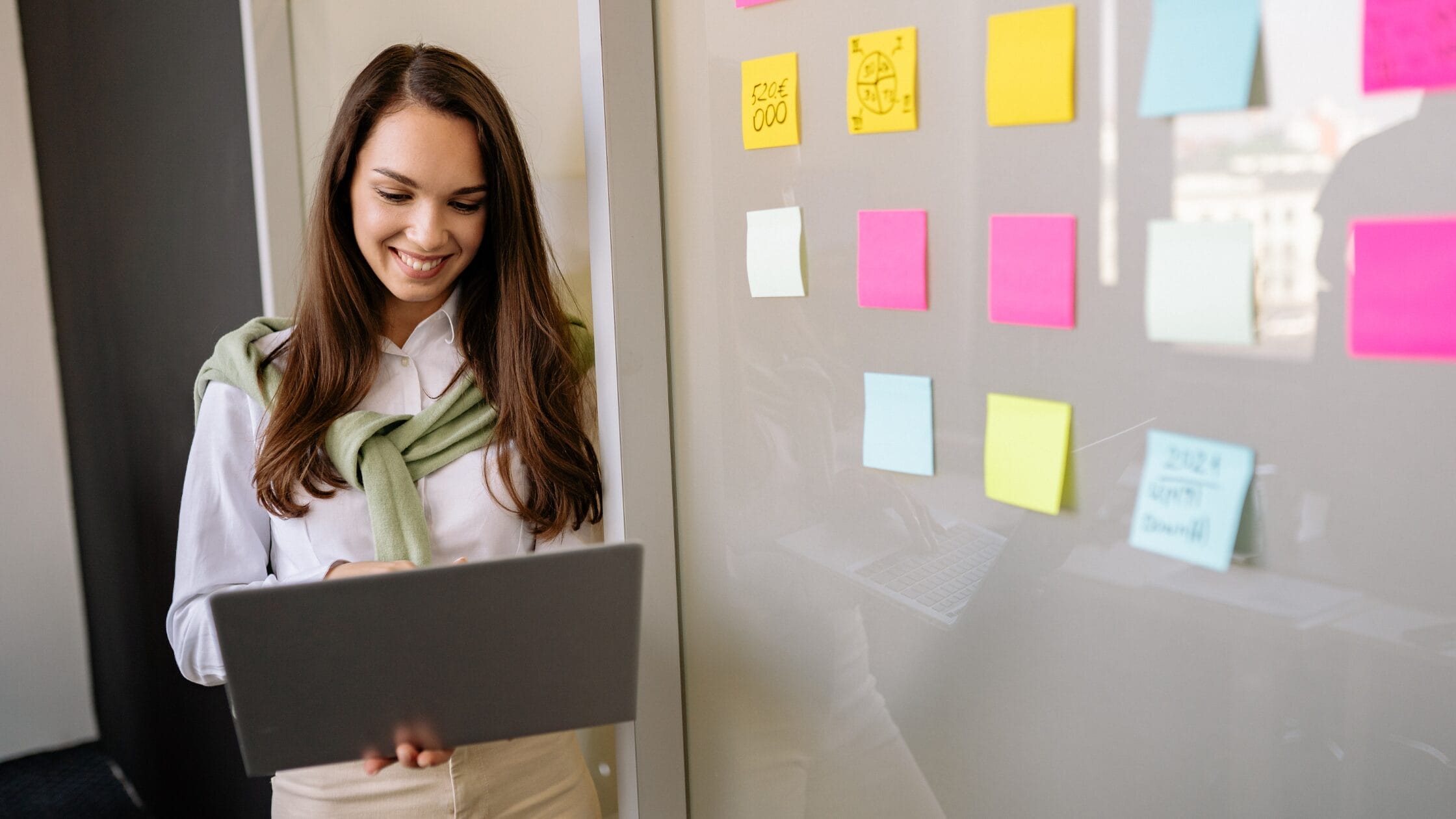 Professional woman reviewing ideas on a glass board with colourful sticky notes while working on a laptop, representing innovation and strategic thinking for the blog “Fostering Innovation While Managing Risk” by Xseed Lead.